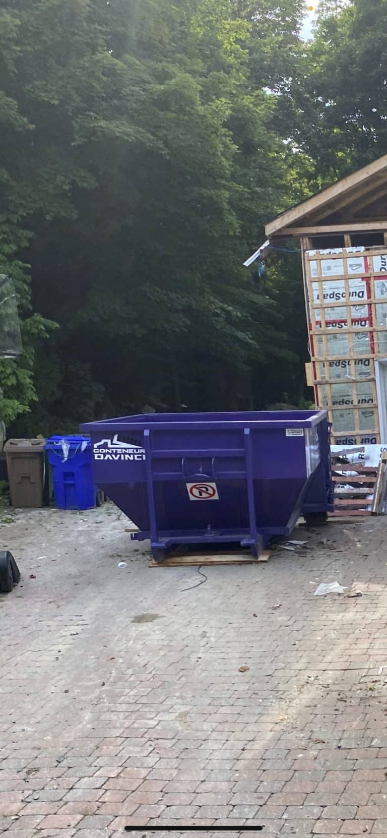 Large purple construction dumpster with Conteneur DaVinci logo on a stone driveway near a partially constructed wooden building surrounded by dense green trees
