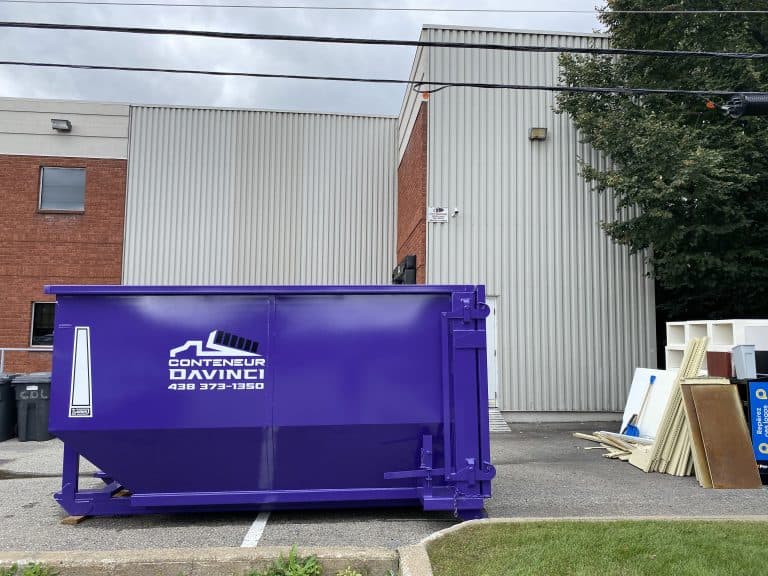 Large purple Davinci container dumpster placed outdoors next to a building with a tree and assorted discarded materials nearby