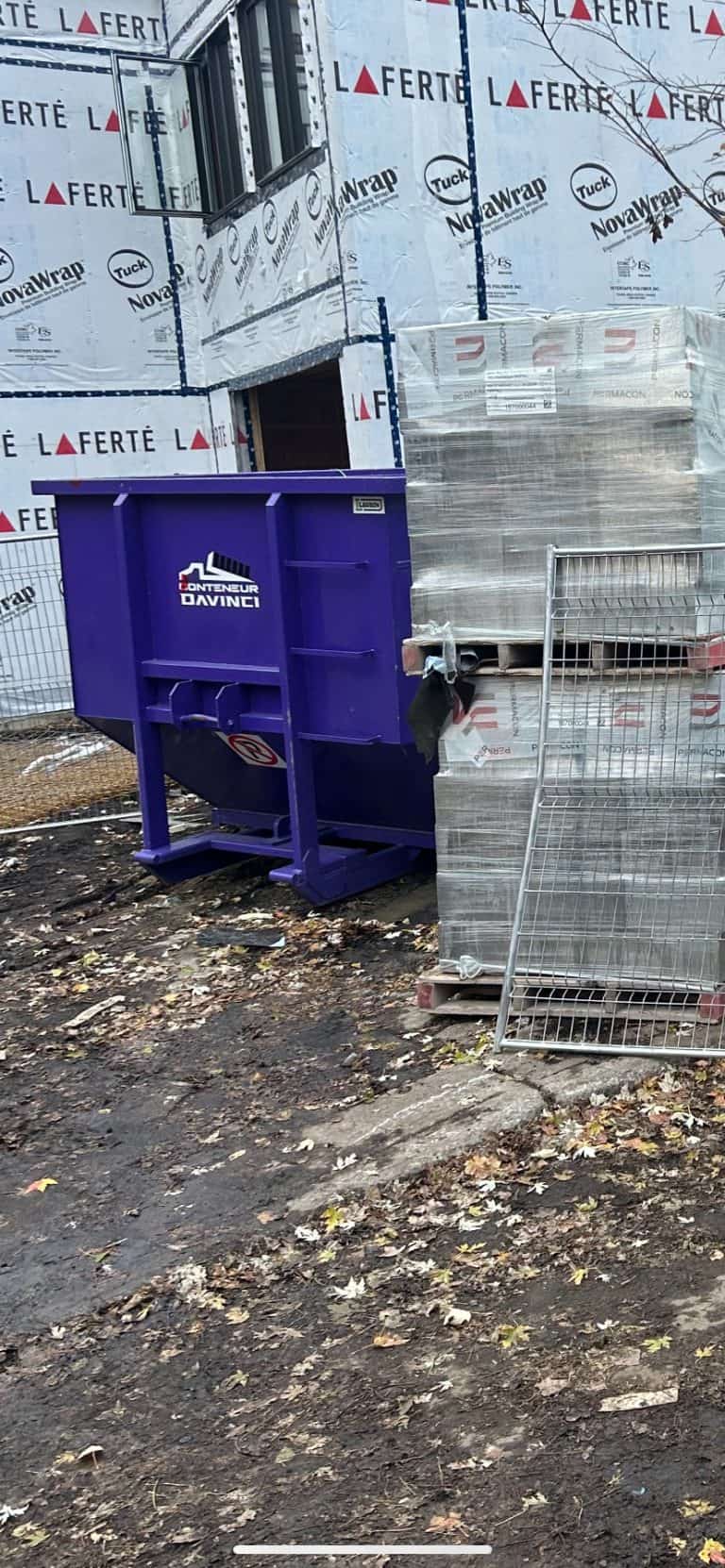 Construction site with a purple Conteneur Davinci dumpster, building wrapped in La Ferte and NovaWrap insulation, and stacked pallets with packaging materials