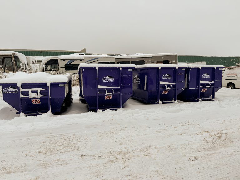 Snow-covered blue Conteneur Davinci dumpsters lined up in a snowy parking lot with RVs and a white van in the background under overcast sky