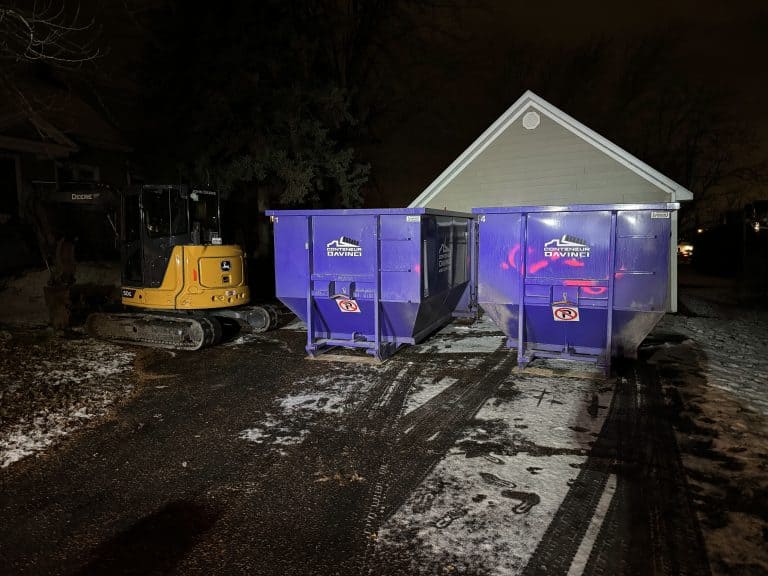 Yellow John Deere excavator parked beside two large purple Conteneur Davinci dumpsters at night with a snowy ground and a house in the background