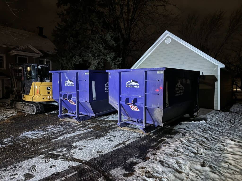 Nighttime view of a yellow John Deere excavator and two large blue Conteneur Davinci dumpsters outside near a small white shed with snow and tire tracks on the ground