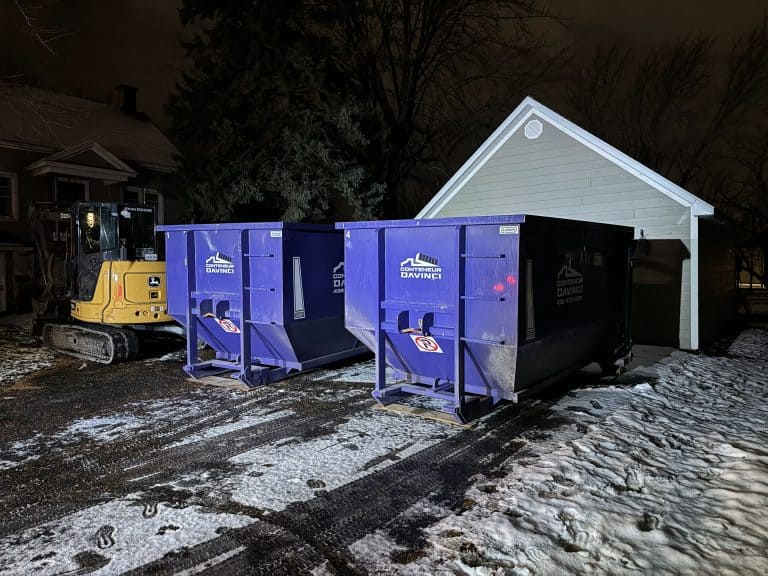 Nighttime view of a yellow John Deere excavator and two large blue Conteneur Davinci dumpsters outside near a small white shed with snow and tire tracks on the ground