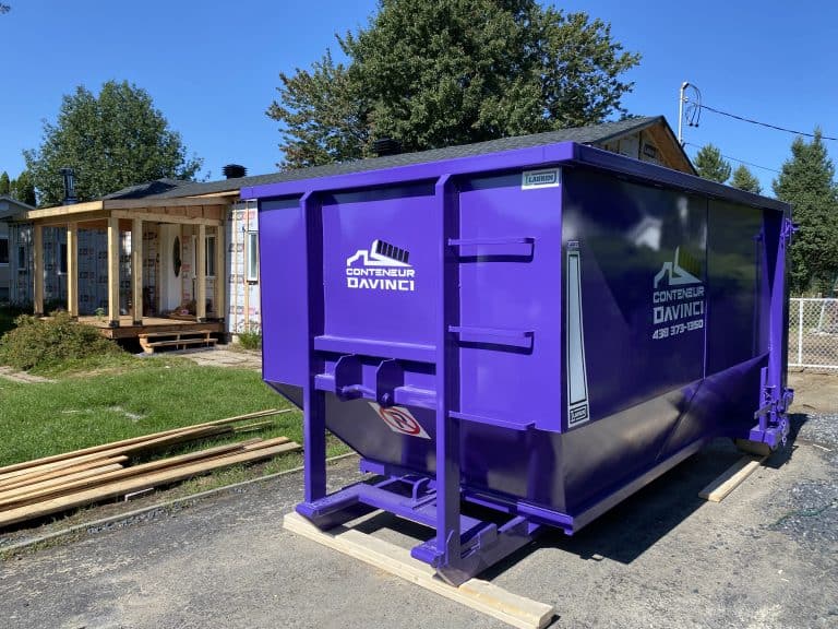 Purple Conteneur Davinci dumpster container at a residential construction site with wooden framing and siding materials.